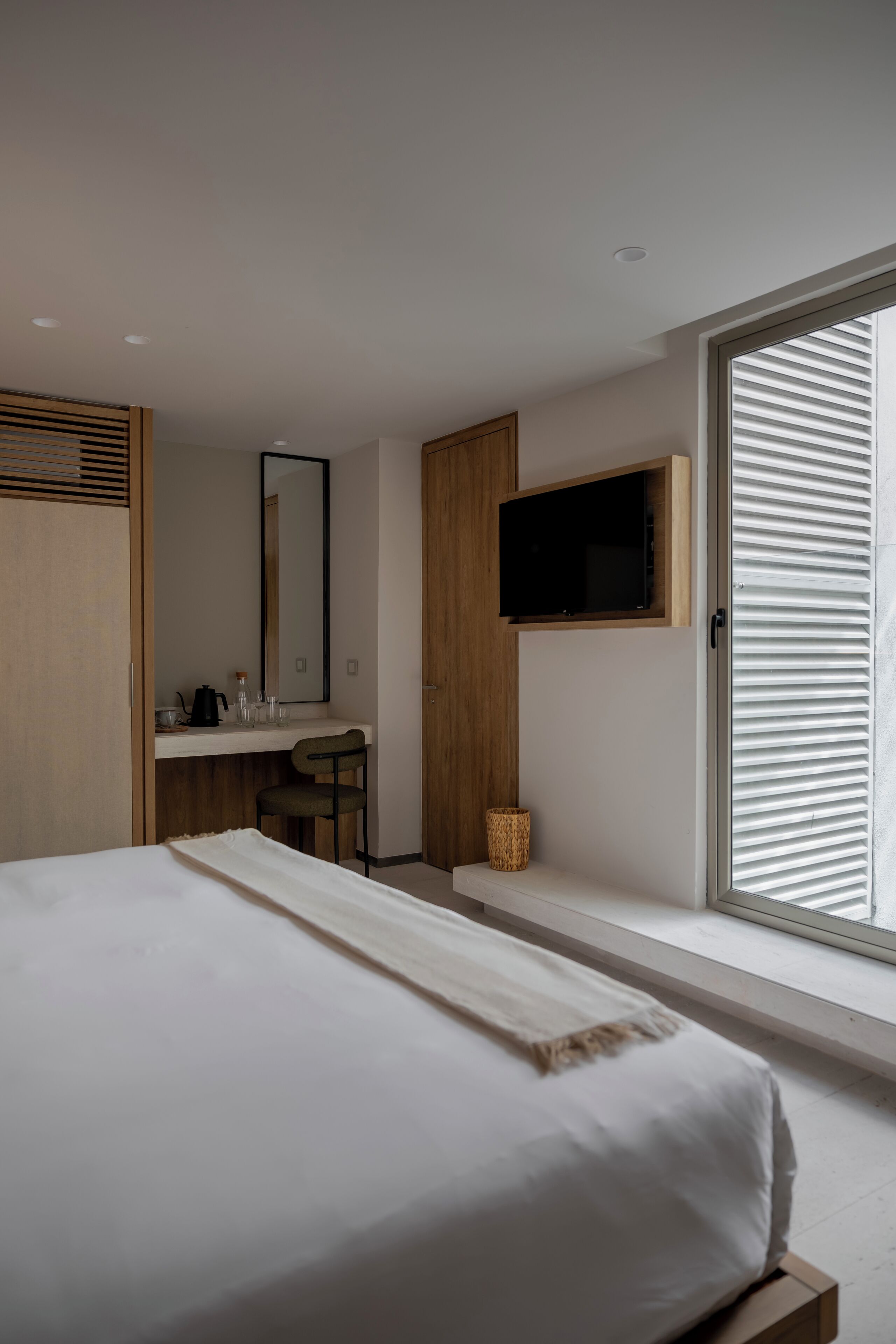 Quarto com cama, escrivaninha, cadeira, televisão e janela com persianas.