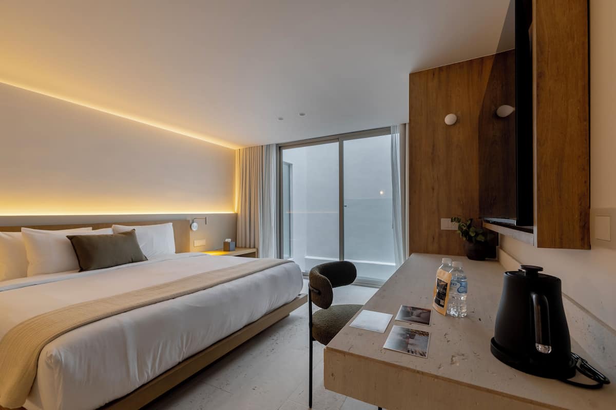 A modern hotel room with a large bed, a desk with a kettle, and a view of the city.