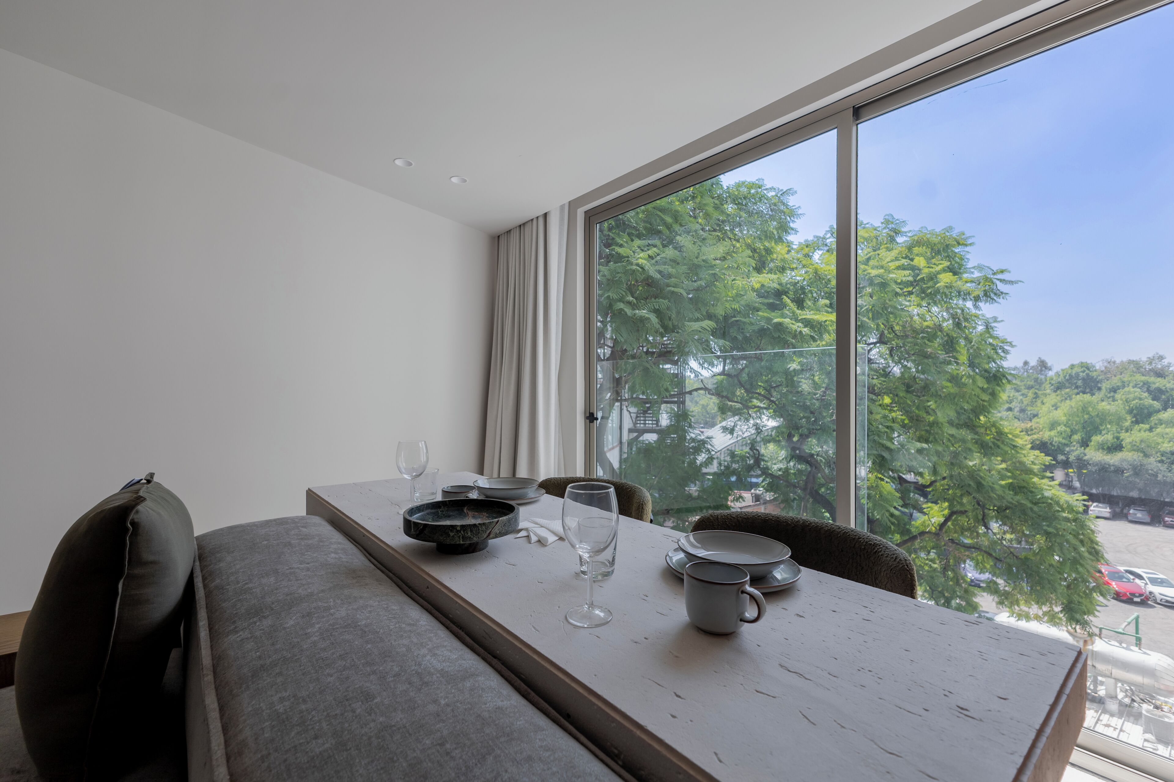 A dining area with a table set for two, a window with a view of trees and parked cars, and a modern interior design.
