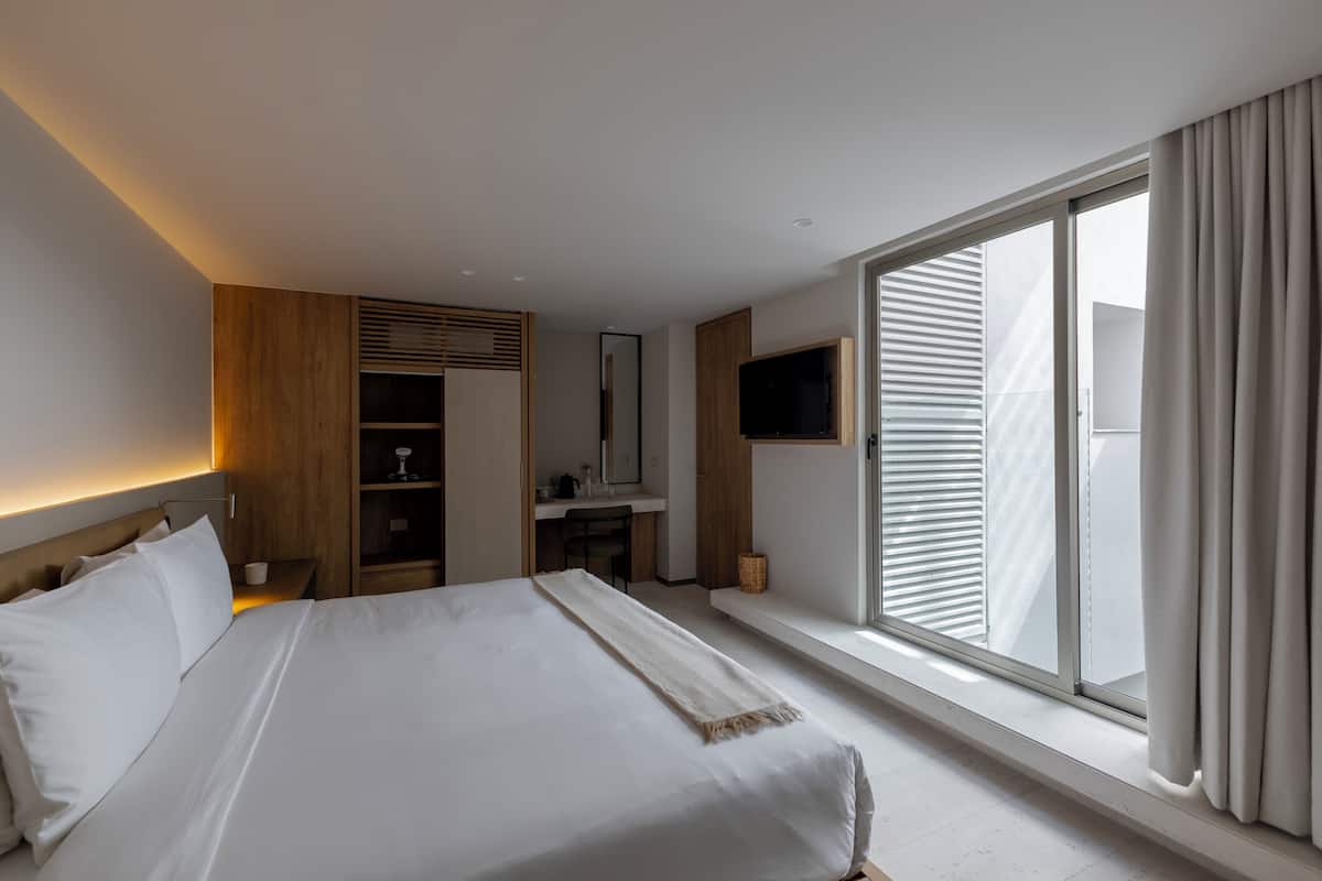 A modern hotel room with a large bed, a desk, a TV, and a window with blinds.