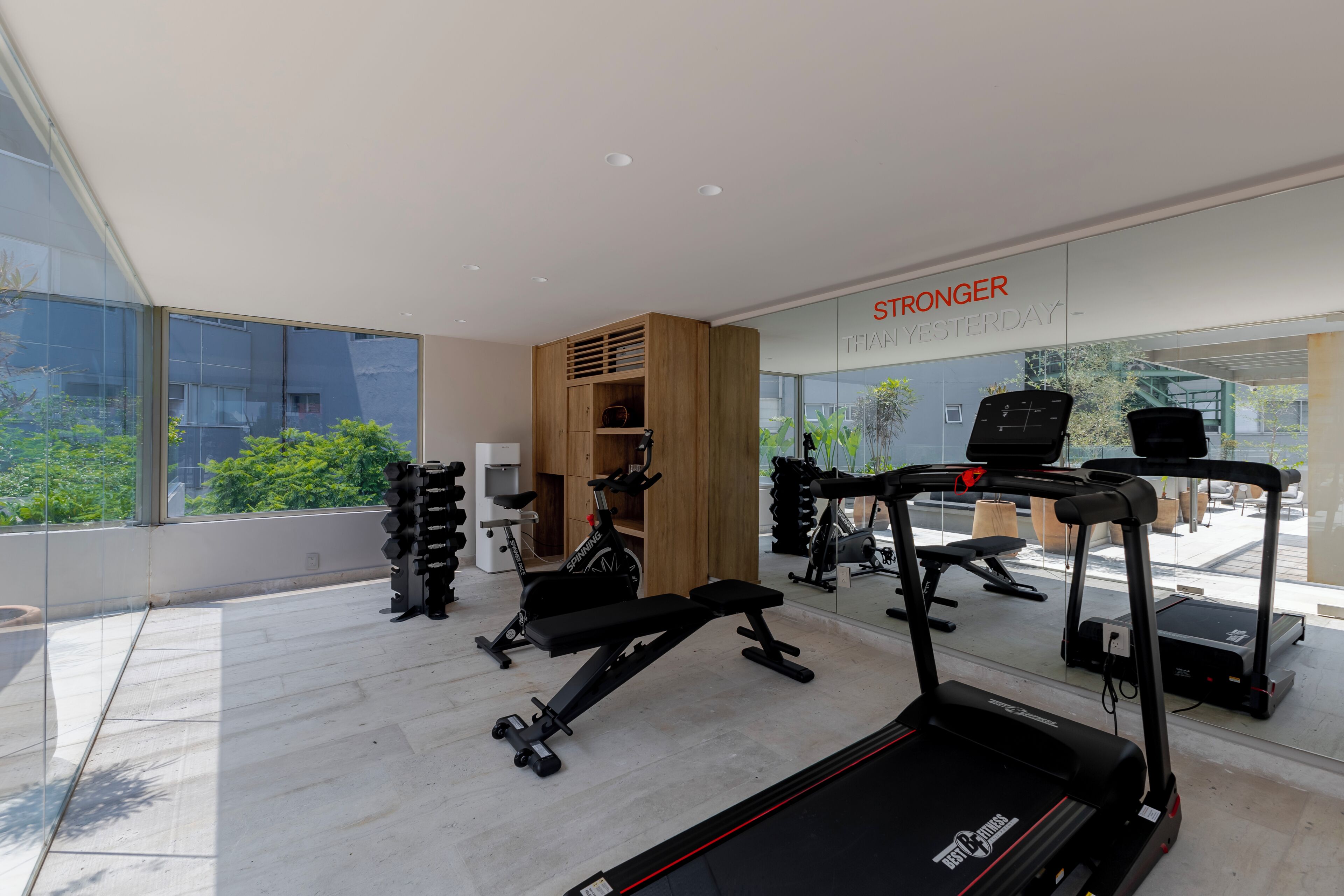 A modern gym with various exercise equipment, large windows, and a view of greenery outside.