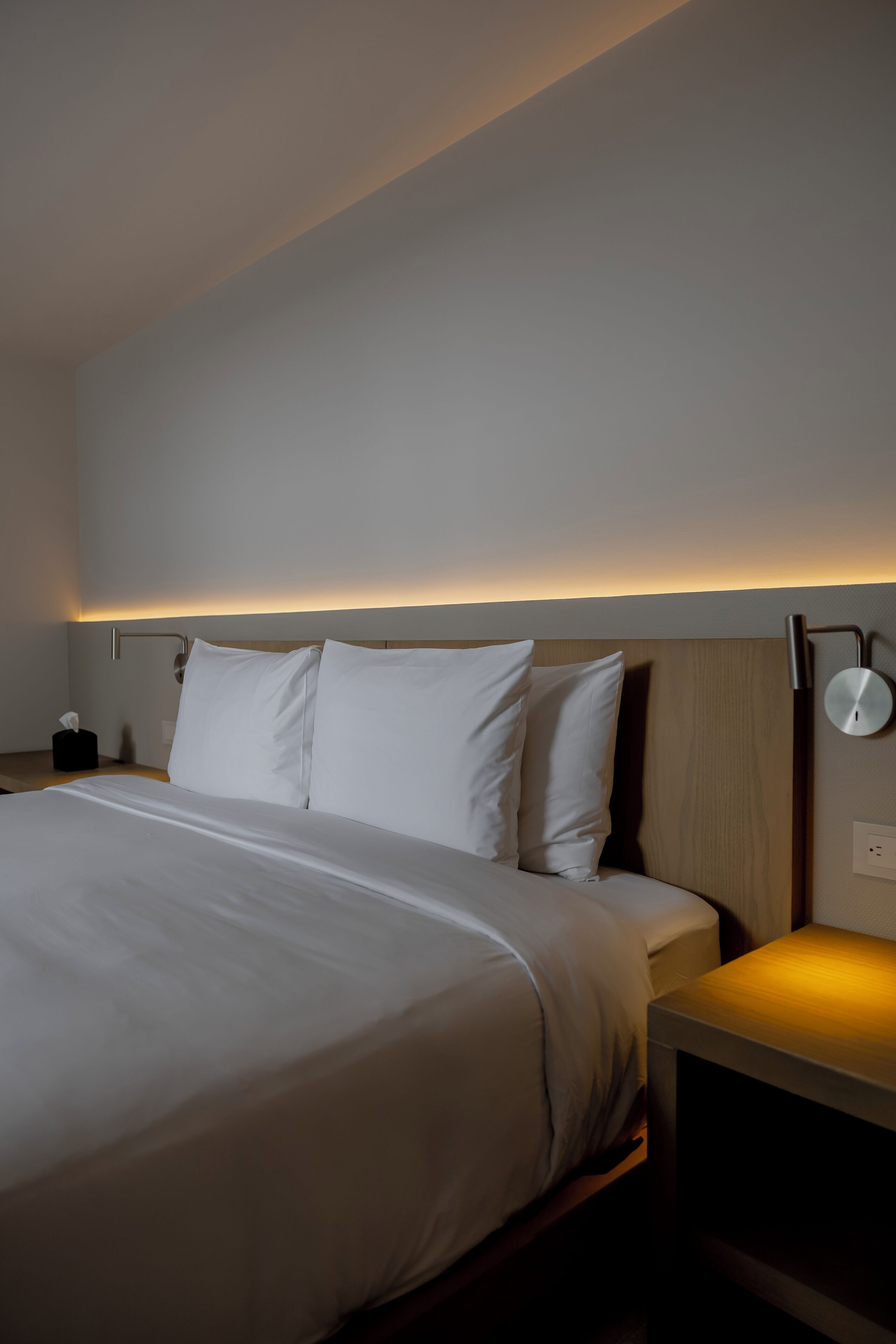 A hotel room with a neatly made bed, a bedside table with a lamp, and a wall-mounted light fixture.