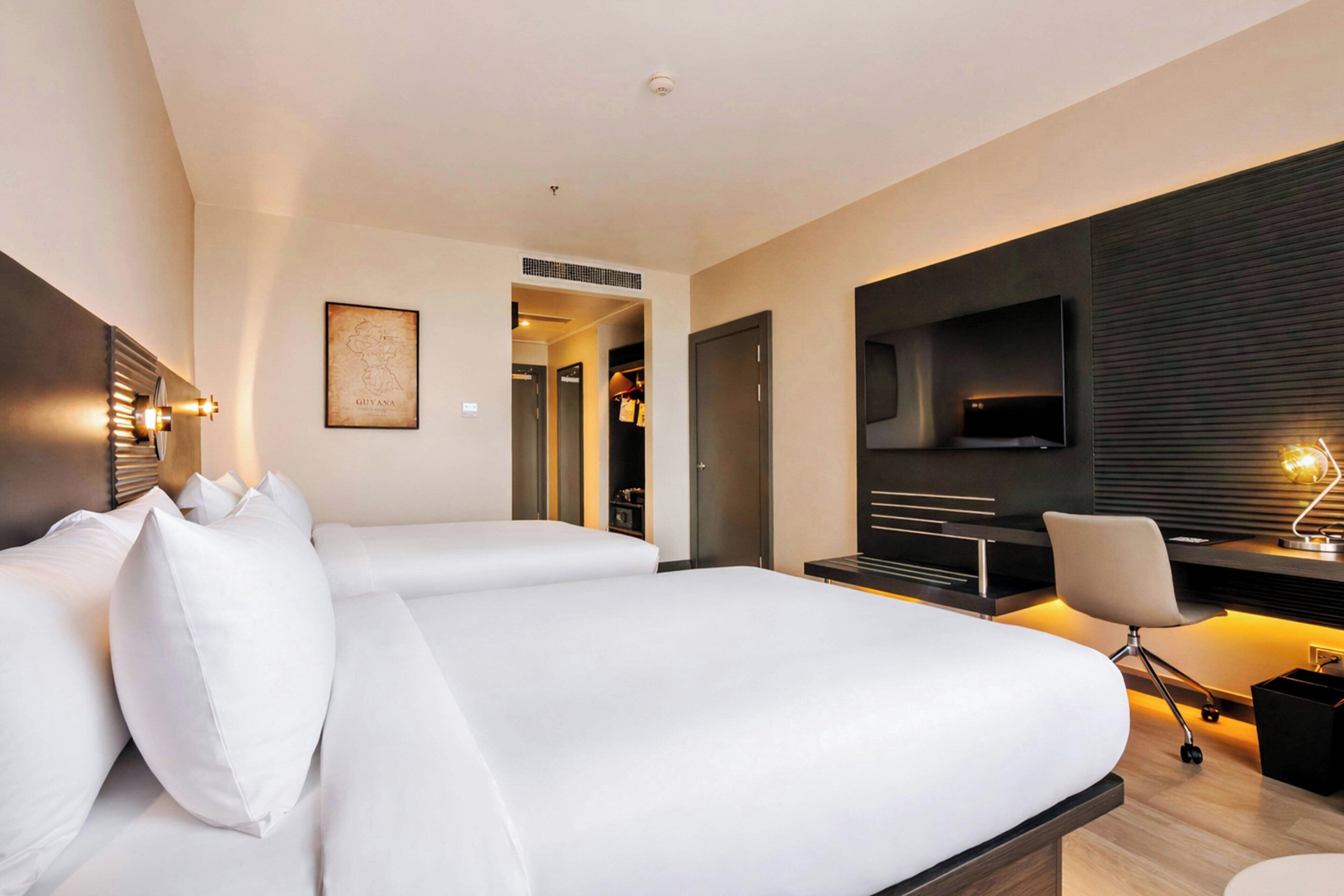A modern hotel room with two beds, a desk, a chair, a TV, and a map on the wall.