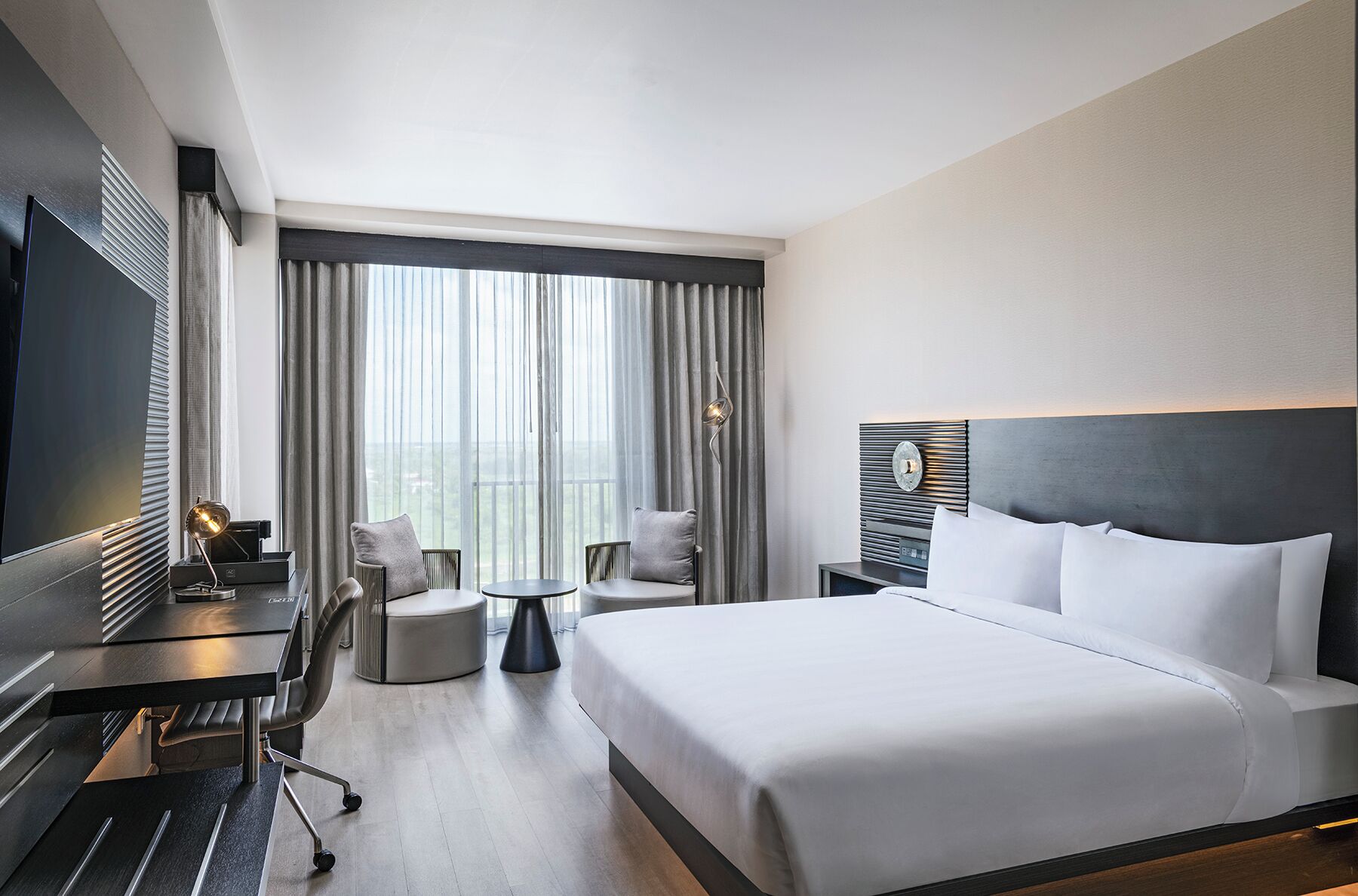 Une chambre d’hôtel avec un grand lit, un bureau avec un ordinateur, une chaise, une petite table et une grande fenêtre avec des rideaux.