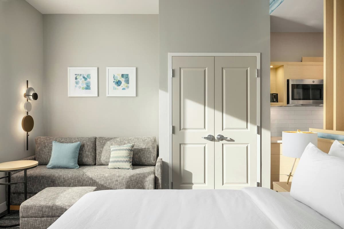 A modern hotel room with a bed, a sofa, a small table, and a door leading to a kitchen area.