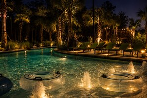 Un área de piscina nocturna con agua cristalina, sillas de descanso y palmeras.