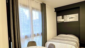 Een hotelkamer met een ronde tafel, twee stoelen, een bed met een beddeken met patroon, een kunstwerk aan de muur en een raam met gordijnen.