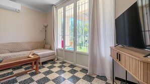A living room with a sofa, a coffee table, a TV on a wooden stand, and a large window with curtains. - Awesome home in Fontvieille (Fontvieille)