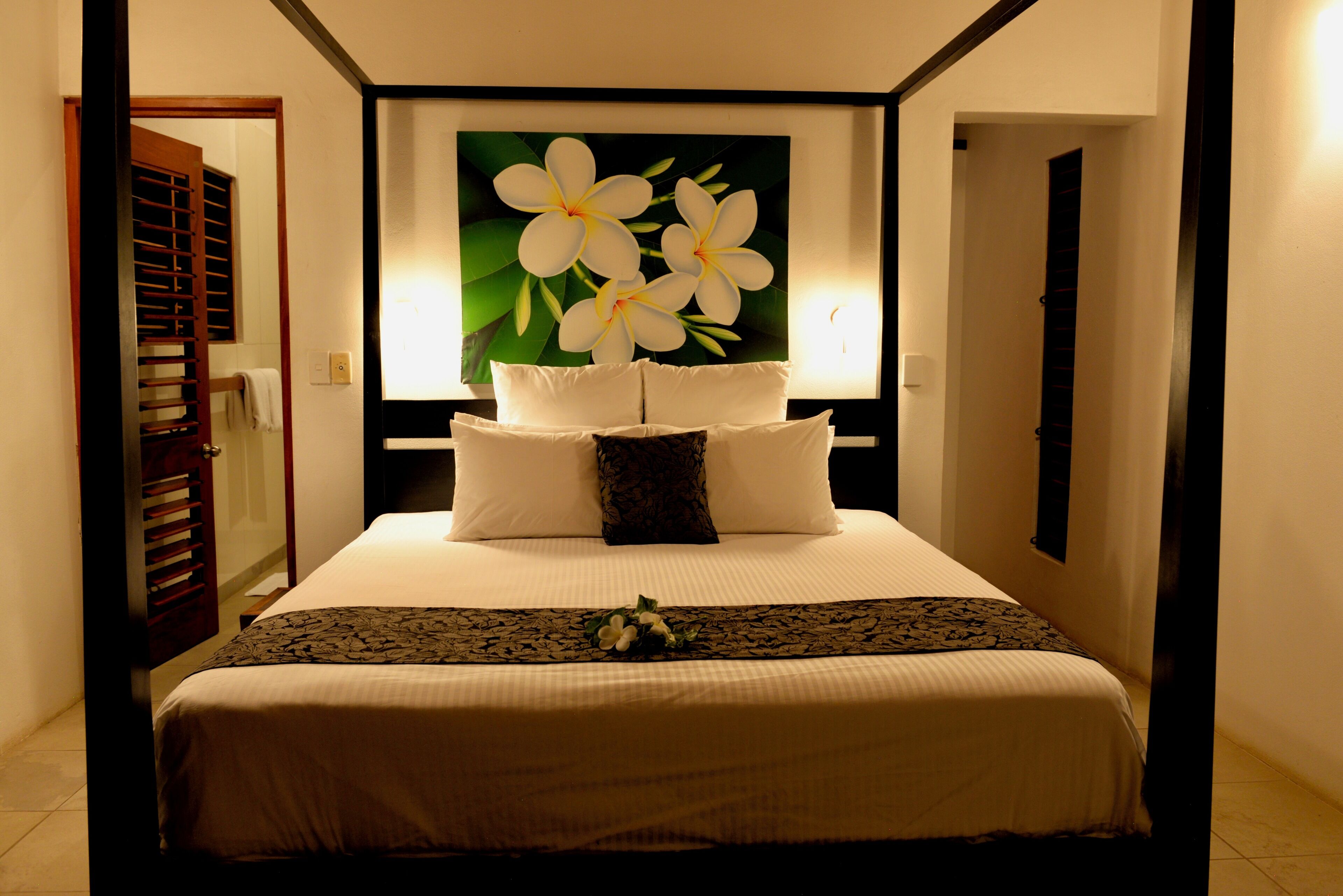 A neatly made bed with white linens and a floral pillow, a large floral artwork above the bed, and a dark wooden headboard.