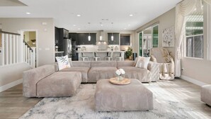 A modern living room with a large sectional sofa, a round ottoman, and a dining area in the background. - Disneyland & Chill w/ Theater/Gameroom/HotTub AN18 (Anaheim)