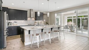 A modern kitchen with dark cabinets, a large island with bar stools, and a dining area with a glass door leading outside. - Disneyland & Chill w/ Theater/Gameroom/HotTub AN18 (Anaheim)