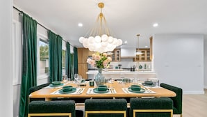A dining area with a wooden table set for six, green upholstered chairs, a vase with flowers, and a modern chandelier. - Foosball, Hot Tub, Big Backyard, Disneyland, AN12 (Anaheim)