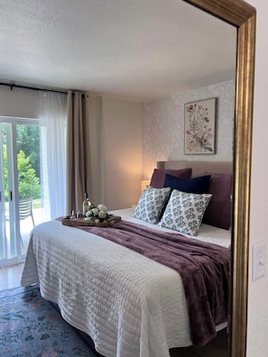 Quarto com cama, mesa de cabeceira, obra de arte floral emoldurada e vista através de uma porta de vidro.