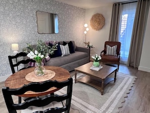 Uma sala de estar com sofá, duas poltronas, mesa de centro, mesa de jantar com cadeiras, abajur e um vaso com flores.