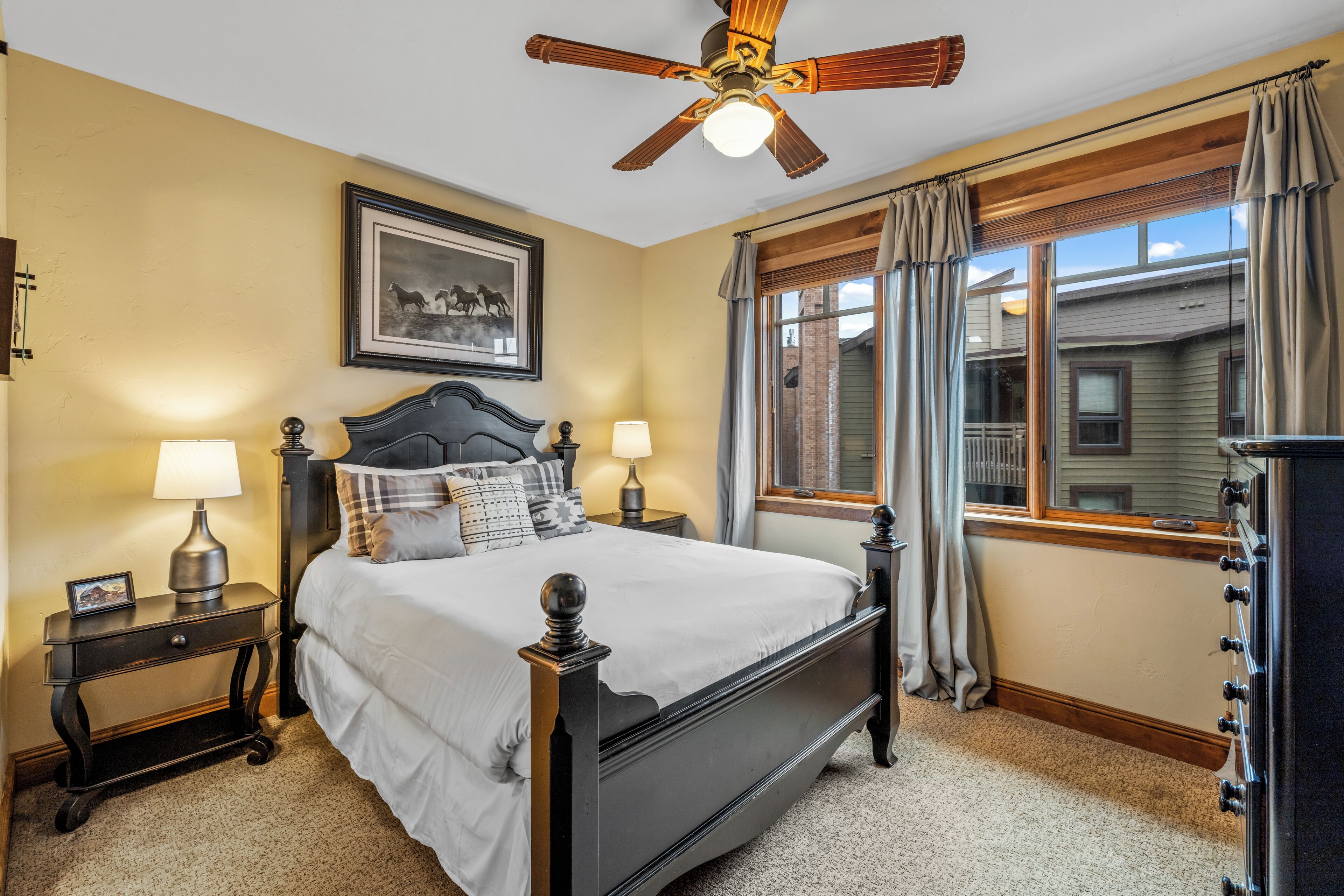 Une chambre avec un grand lit, deux tables de chevet avec des lampes, un ventilateur de plafond, une fenêtre avec des rideaux et un tableau encadré au mur.