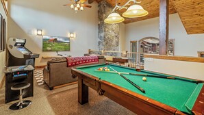 Una mesa de billar con superficie de fieltro verde, una vara de billar y una bola blanca. Hay una zona de bar con una mesa de billar, un taburete y una máquina. Un televisor está montado en la pared, y se ve una pared de piedra.