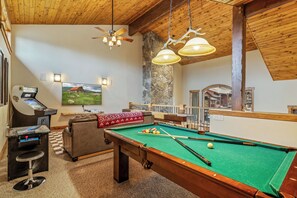 A pool table with a green felt surface, a pool cue, and a cue ball. There is a bar area with a pool table, a stool, and a machine. A television is mounted on the wall, and a stone wall is visible. - Cimarron #14 - walk to base, winter ski shuttle, private deck spa, Mountain views! (Steamboat Springs)