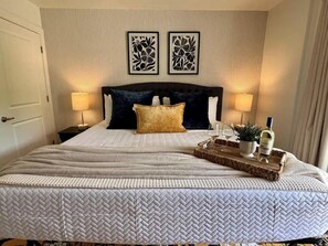Un lit avec une couvre-lit à motifs, deux cadres photo accrochés au mur, des lampes de chevet et un plateau avec une bouteille et des verres.