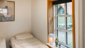 A bedroom with two beds, a window with a view of a house and outdoor seating, and a bedside lamp.