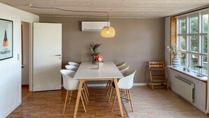 A dining area with a white table, six white chairs, a hanging light fixture, a vase with flowers, and a window with a view of greenery.