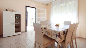 A dining area with a table covered in a coffee-themed tablecloth, surrounded by chairs. A white cabinet with shelves is visible to the left. - Apartment 2411/167402 (Istria - Liznjan), 1500m from the beach (Liznjan)