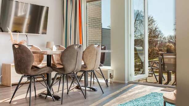 A modern dining area with a table and four chairs, a flat-screen TV mounted on the wall, and a view of the outdoors through a glass door.