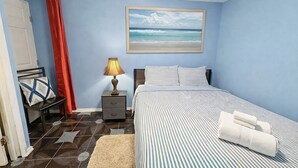 Un dormitorio con cama, mesita de noche, lámpara, silla, cortina roja y un cuadro de una playa.