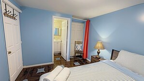 Habitación de hotel con cama, mesita de noche, lámpara, silla y baño visible a través de una puerta abierta.