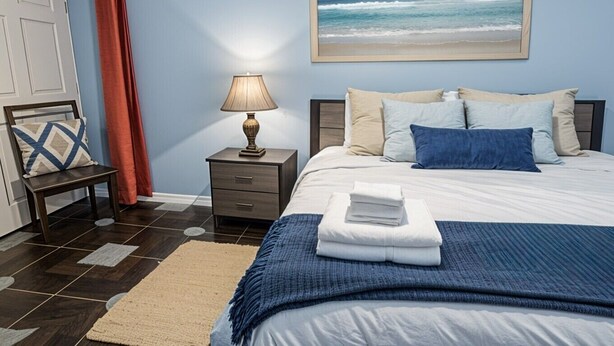 Un dormitorio con cama, mesita de noche, lámpara, silla, puerta y un cuadro del océano.