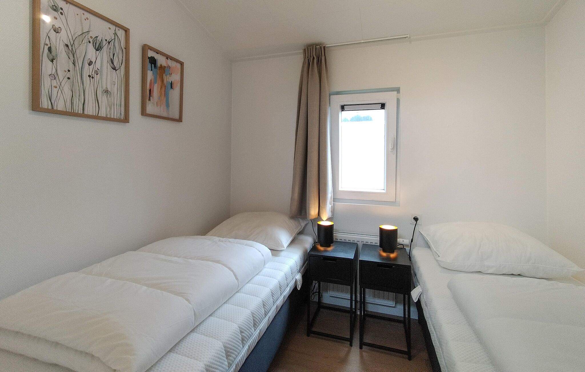 A small, neatly arranged room with two single beds, a small table with two lamps, and a window with curtains.