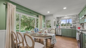Un comedor con una mesa dispuesta para cuatro personas, rodeada de sillas, y una cocina con armarios verdes y una ventana con vistas a la vegetación.