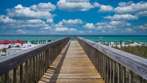 Eine Holzstege führt zu einem Strand mit Sonnenschirmen und Liegestühlen.