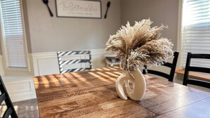 Une table à manger ornée d’un vase de fleurs séchées, un cadre décoratif accroché au mur et une fenêtre avec des stores.