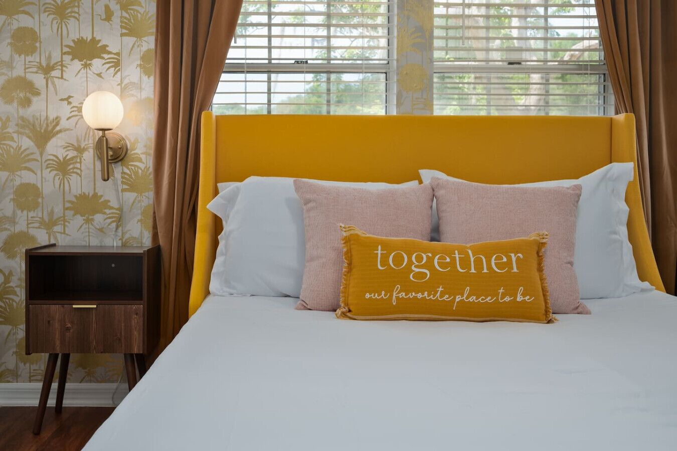 A bed with a yellow headboard, white and pink pillows, and a 'together our favorite place to be' pillow.
