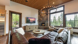 A spacious living room with a stone fireplace, leather sofas, and large windows offering a view of trees. - Monthly Lease - Ambiance at Martis Camp by Tahoe Getaways - 4BR w/ Hot Tub & Bonus Room (Truckee)