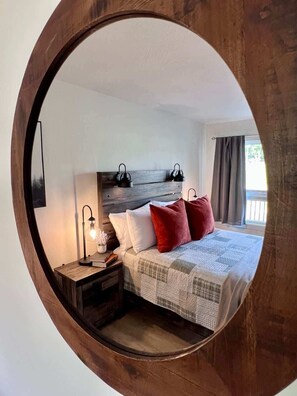 A bedroom with a wooden headboard, a bed with white and red pillows, a bedside table with a lamp, and a view of the outdoors through a window.