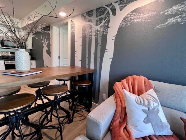 Un comedor con mesa y sillas de madera, un jarrón con ramas y un mural en la pared con motivos de árboles.