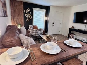 Un coin repas avec une table en bois dressée pour quatre personnes, un canapé, un fauteuil et un téléviseur fixé au mur.