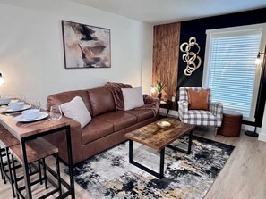 Una sala de estar moderna con un sofá de cuero marrón, una mesa de centro de madera, un sillón a cuadros y un comedor con una mesa y sillas de madera.