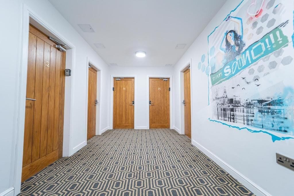 A hallway with patterned carpet, wooden doors numbered 12 to 15, and a wall mural with a cityscape and the word "SOUND".