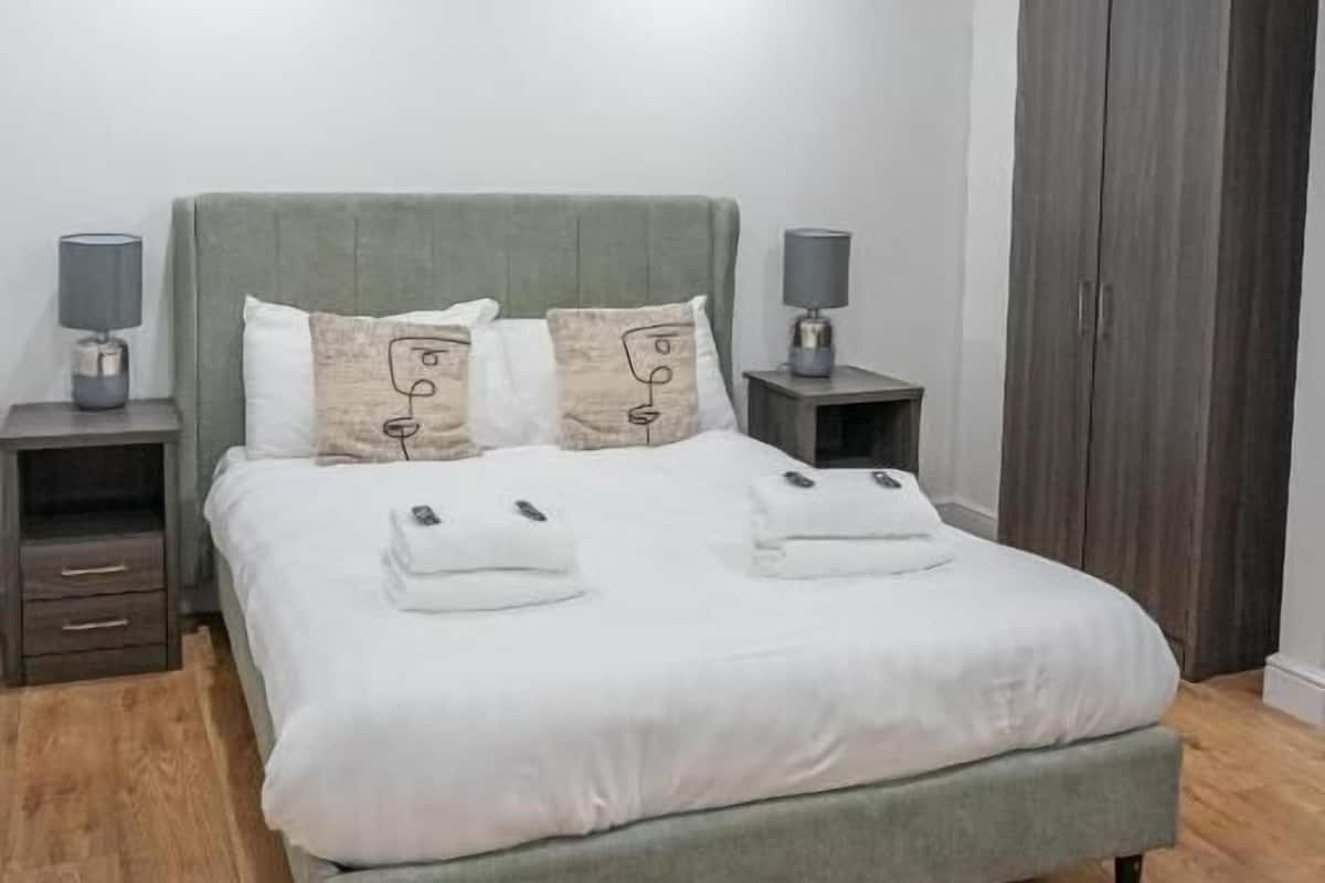 A modern bedroom with a grey upholstered bed, two bedside tables with lamps, a wardrobe, and a wooden floor.