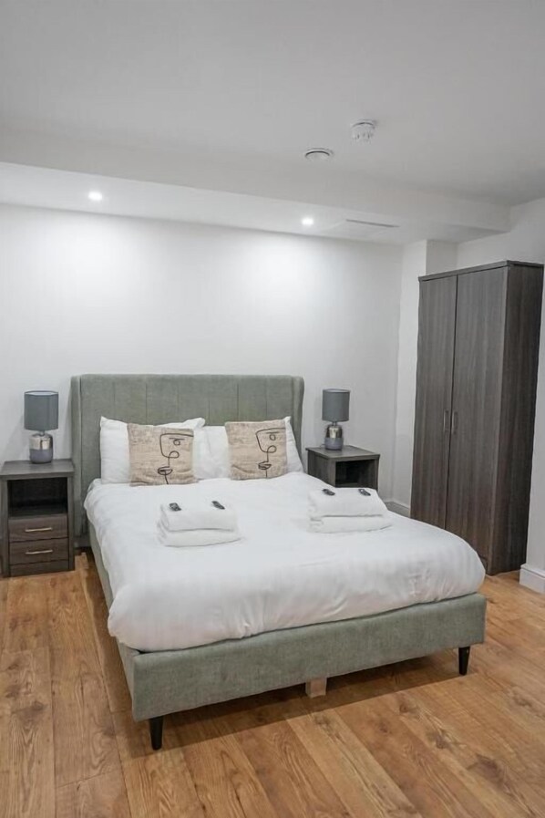 A modern bedroom with a grey upholstered bed, two bedside tables with lamps, a wardrobe, and a wooden floor.