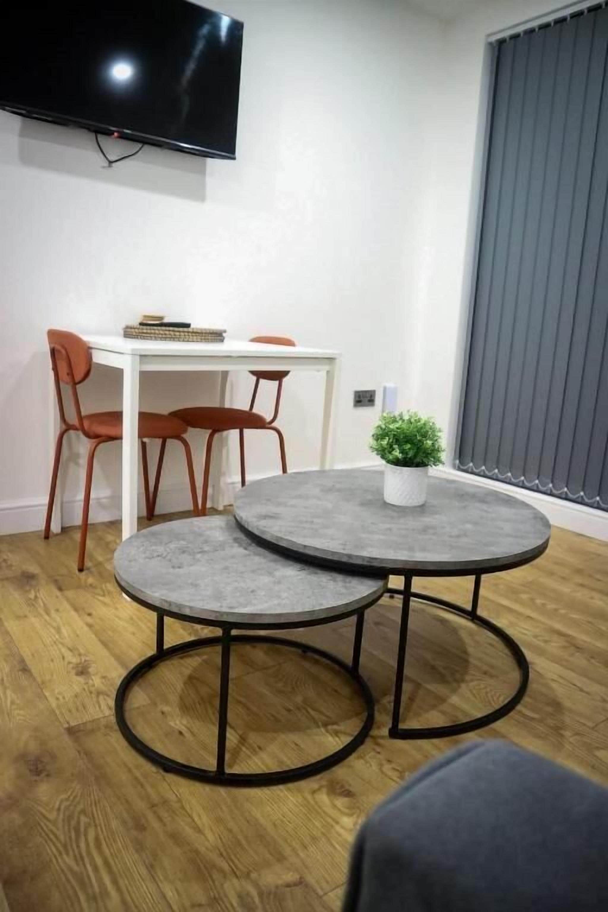 A modern living room with a round grey coffee table, a white desk with two orange chairs, a flat-screen TV mounted on the wall, and a potted plant on the coffee table.