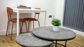 A modern living room with a round grey coffee table, a white desk with two orange chairs, a flat-screen TV mounted on the wall, and a potted plant on the coffee table.