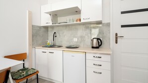 Una cucina compatta con mobili bianchi, un lavandino, un bollitore e una sedia con cuscino verde.