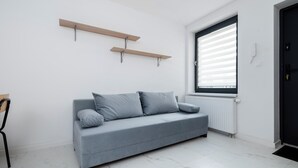 Una habitación moderna y minimalista con un sofá gris, dos estantes de madera, un radiador y una ventana con persianas.