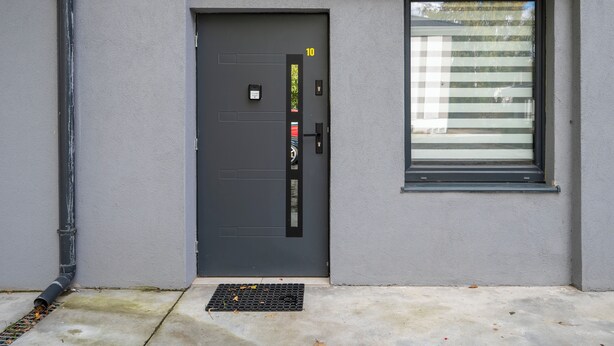 Una puerta gris moderna con cerradura electrónica, un pequeño ventanal con persianas horizontales y una rejilla metálica en la parte inferior.