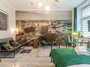 A modern hotel room with a cityscape mural, a bed with green bedding, a desk with a lamp, and a seating area with a sofa and chairs. - Strzelecka Central Studio (Katowice)