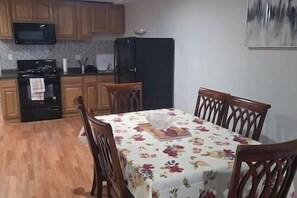 A compact kitchen with wooden cabinets, a black refrigerator, and a microwave. A dining table with floral tablecloth and wooden chairs is centered in the room.