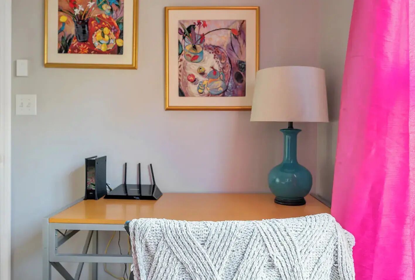 A room with a wooden desk, a teal lamp, and two framed paintings on the wall.